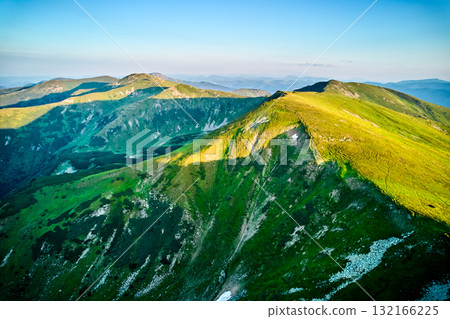 Aerial view of rugged mountain range with lush green slopes at sunset. Landscape showcases beauty of untouched nature with distant peaks and valleys bathed in soft sunlight. Carpathians, Chornogora. 132166225