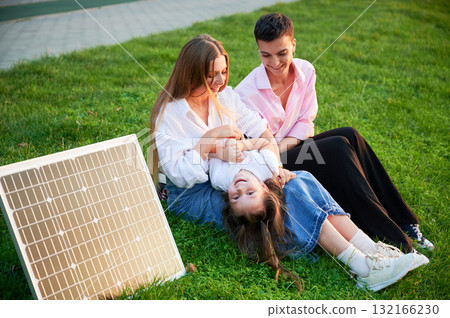 Young women using solar panel for charging, sit on grassy lawn, smiling and interacting with small child lying playfully on woman lap. Girls casually dressed, enjoying sunny, relaxed moment outdoors. 132166230