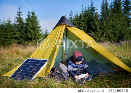 Young child plays with smartphone while charging with photovoltaic solar panel near tourist tent in summer. Integration of renewable energy in outdoor camping activities. Young child plays with smartphone while charging with photovoltaic solar panel near tourist tent in summer. Integration of renewable energy in outdoor camping activities. 132166236