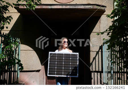 Woman holding solar panel for charging, stands in front of old building's entrance. Smiling girl wears sunglasses, white top, and blue jeans. Scene framed by green foliage and iron fencing. 132166241
