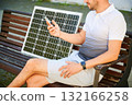 Cropped view of man using smartphone connected to photovoltaic solar panel. Integration of sustainable renewable energy into everyday life, showing practical use of solar power for charging devices. 132166258