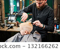 Man barber cutting little boy's hair using comb and scissors. Child getting haircut from adult male, likely barber. Professional hairdresser and cute client at modern barbershop. 132166262