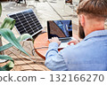 Man works on laptop at outdoor round table, with glass of iced coffee. Solar panel nearby, emphasizing sustainable, modern eco-friendly workspace that combines technology and sustainability. 132166270