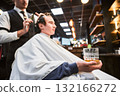 Close-up photo of young client holding glass of whiskey. Male customer relaxing while getting new haircut. Professional hairdresser in fancy outfit cutting client's hair. 132166272