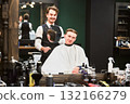 Happy hairdresser showing new haircut to his client. Barber using round mirror to show back view of customer's head. Young male satisfied with new hairstyle. 132166279