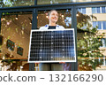 Woman holding solar panel for charging, stands outdoors, smiling in urban setting. Concept of green energy generation. 132166290