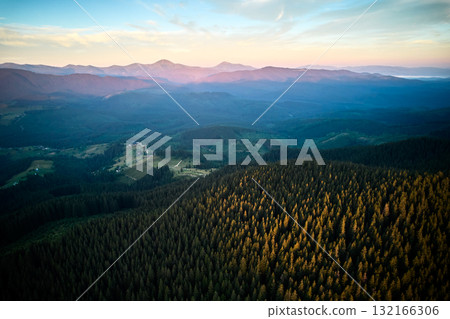 Aerial view of sunlit forested mountain with long shadows casting across vibrant green trees. Horizon stretches to distant mountains under clear blue sky, creating serene and expansive landscape. 132166306