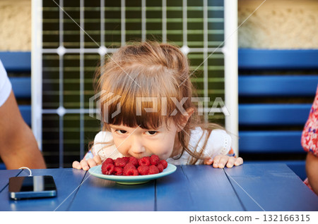 Lovely moment of child eating berries without hands. Daughter having fun with her parents. Little girl eating raspberries on background of solar battery. 132166315