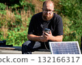 Bearded man using smartphone connected to photovoltaic solar panel. Integration of sustainable renewable energy into everyday life, demonstrating practical use of solar power for charging devices. 132166317