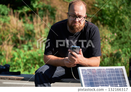 Bearded man using smartphone connected to photovoltaic solar panel. Integration of sustainable renewable energy into everyday life, demonstrating practical use of solar power for charging devices. 132166317