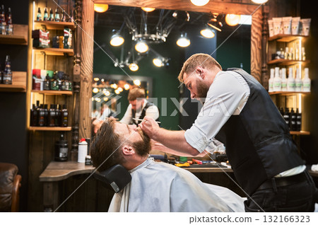 Handsome man getting his beard done. Male customer relaxing while professional hairdresser cutting his beard. Experienced barber using comb and scissors. Handsome man getting his beard done. Male customer relaxing while professional hairdresser cutting his beard. Experienced barber using comb and scissors. 132166323