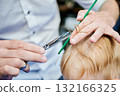 Man barber cutting little boy's hair using comb and scissors. Child getting haircut from adult male, likely barber. Professional hairdresser and cute client at modern barbershop. 132166325
