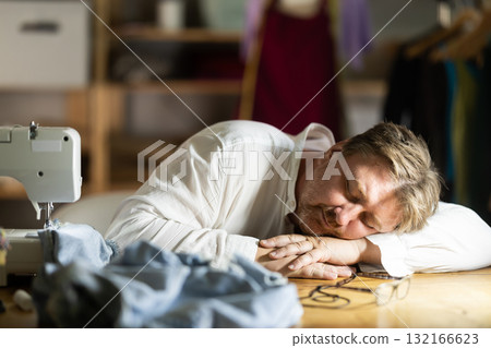 Late in evening, tired man tailor dozed off next to sewing machine. Late in evening, tired man tailor dozed off next to sewing machine. 132166623
