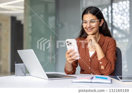 Happy businesswoman smiling while using smartphone and working on laptop at her desk in a modern office, enjoying the blend of technology and communication in her career Happy businesswoman smiling while using smartphone and working on laptop at her desk in a modern office, enjoying the blend of technology and communication in her career 132169189