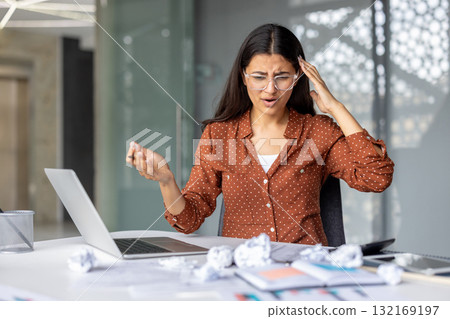 Stressed businesswoman is having headache while working on difficult task on laptop in office, surrounded by crumpled paper, showing frustration and burnout 132169197