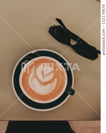 Coffee art and sunglasses on a table. A latte with beautiful coffee art sits on a saucer next to a pair of black sunglasses on a table, top-down view. Coffee art and sunglasses on a table. A latte with beautiful coffee art sits on a saucer next to a pair of black sunglasses on a table, top-down view. 132170678