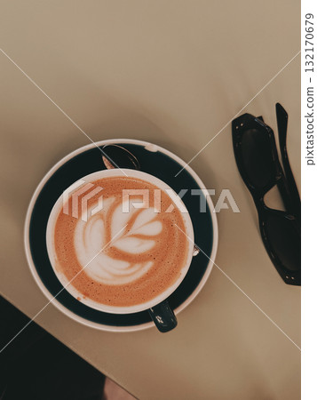 Coffee art and sunglasses on a table. A latte with beautiful coffee art sits on a saucer next to a pair of black sunglasses on a table, top-down view. 132170679