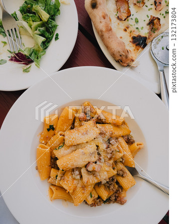 Rigatoni pasta with bolognese sauce, grated cheese and parsley served on white plate on restaurant table with pizza and green salad in background. Flatlay of pizza, rigatoni pasta and green salad 132173804