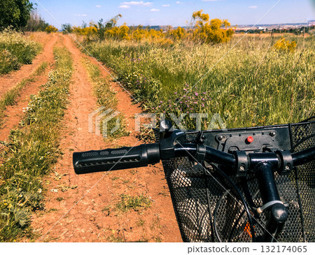 A bicycle handlebar and basket on a dirt road through a field of tall grass. The image captures a bicycle's handlebars and basket, with a dirt road leading into a field of tall grass under bright sky. A bicycle handlebar and basket on a dirt road through a field of tall grass. The image captures a bicycle's handlebars and basket, with a dirt road leading into a field of tall grass under bright sky. 132174065