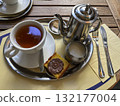 Cozy breakfast scene with a cup of tea, metal teapot, milk jug, and a small cake served on a tray in a cafe setting, evoking a warm and relaxing morning atmosphere 132177004