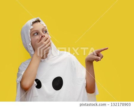 Smiling boy dressed as ghost is pointing upward, standing in front of vibrant yellow background Halloween. Smiling boy dressed as ghost is pointing upward, standing in front of vibrant yellow background Halloween. 132178903