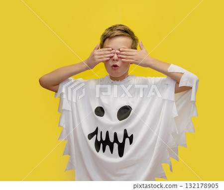 Smiling boy dressed as ghost is pointing upward, standing in front of vibrant yellow background, likely themed for Halloween. Smiling boy dressed as ghost is pointing upward, standing in front of vibrant yellow background, likely themed for Halloween. 132178905