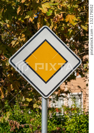 Priority road sign in front of autumn foliage. Yellow diamond-shaped priority road sign stands out against background of autumn trees and sunny day. Concept of traffic regulation, road safety signal Priority road sign in front of autumn foliage. Yellow diamond-shaped priority road sign stands out against background of autumn trees and sunny day. Concept of traffic regulation, road safety signal 132179082