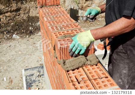 A skilled bricklayer is carefully applying mortar to red bricks, aligning them precisely to build a wall at a construction site A skilled bricklayer is carefully applying mortar to red bricks, aligning them precisely to build a wall at a construction site 132179318