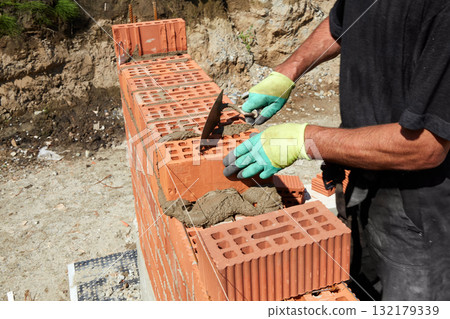 A skilled bricklayer is carefully applying mortar to red bricks, aligning them precisely to build a wall at a construction site 132179339