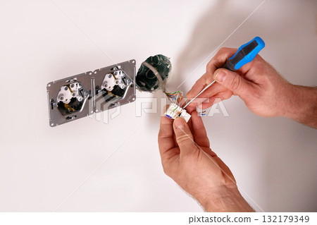 A person is engaged in an electrical repair task at home, carefully working with wires and tools near a wall outlet. A person is engaged in an electrical repair task at home, carefully working with wires and tools near a wall outlet. 132179349