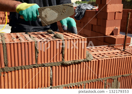A skilled bricklayer is carefully applying mortar to red bricks, aligning them precisely to build a wall at a construction site 132179353