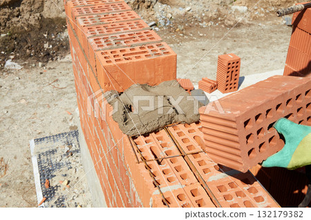 A skilled bricklayer is carefully applying mortar to red bricks, aligning them precisely to build a wall at a construction site A skilled bricklayer is carefully applying mortar to red bricks, aligning them precisely to build a wall at a construction site 132179382