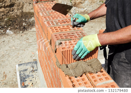 Skilled bricklayer constructs a sturdy wall using red bricks and mortar under bright sunlight in a construction site Skilled bricklayer constructs a sturdy wall using red bricks and mortar under bright sunlight in a construction site 132179480
