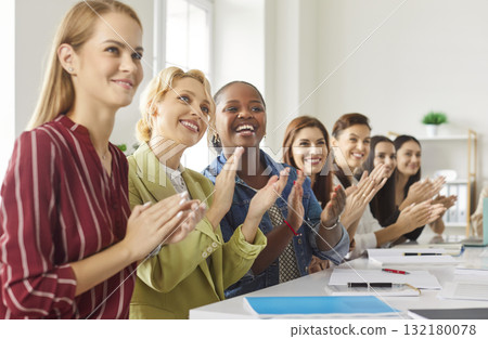 Smiling diverse group of women sitting at desk, clapping hands, supporting presentation or event. 132180078