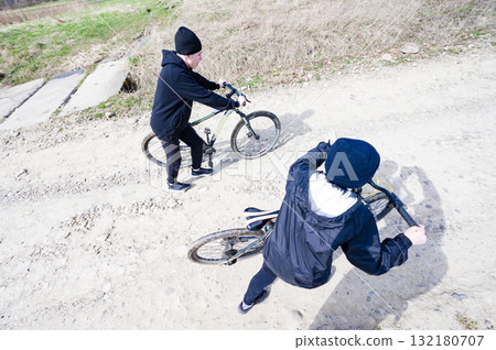 Two cyclists on a dirt road, enjoying a sunny day outdoors. One pushes their bike, the other sits on theirs. 132180707