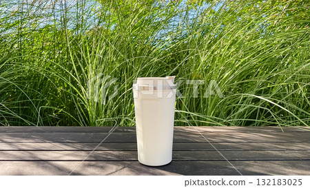 Minimalist white travel mug on wooden deck with lush green reeds. Sustainable coffee/tea in nature 132183025