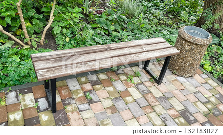 Rustic park bench next to pebble trash can on patterned patio tile among colorful garden flowers 132183027