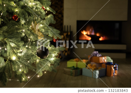Beautifully decorated Christmas tree with lights and ornaments near a glowing fireplace, surrounded by colorful wrapped gifts creating a cozy festive atmosphere. 132183843