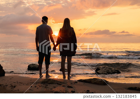 Couple holding hands and watching the sunrise over the sea, standing barefoot on a sandy beach with gentle waves and warm morning light Couple holding hands and watching the sunrise over the sea, standing barefoot on a sandy beach with gentle waves and warm morning light 132183845