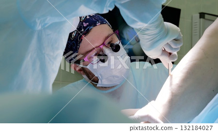 Skilled surgeon wearing magnifying glasses performing delicate surgical procedure, carefully working alongside medical colleague during precision driven, life critical intervention Skilled surgeon wearing magnifying glasses performing delicate surgical procedure, carefully working alongside medical colleague during precision driven, life critical intervention 132184027
