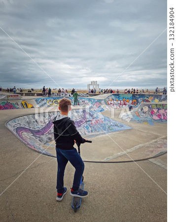 Le Havre, Normandy, France - 29 May, 2025 Rear view of a boy on a scooter looking toward the graffiti-covered skatepark bowl, at Docks Vauban Skatepark near the beach in Le Havre 132184904