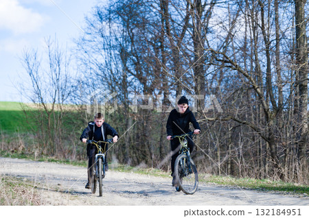 Two boys enjoy a bike ride on a country road, surrounded by trees. A sunny day outdoors. 132184951