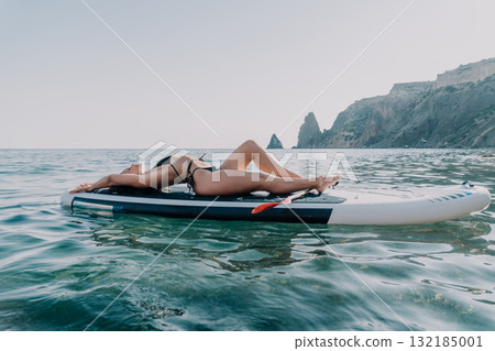 Woman Relaxing Paddleboard Sea - A woman sunbathes on a paddleboard in the ocean near cliffs. Woman Relaxing Paddleboard Sea - A woman sunbathes on a paddleboard in the ocean near cliffs. 132185001