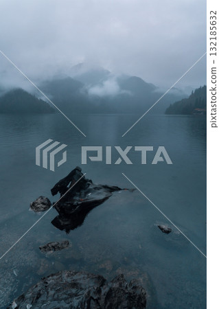 Serene lake landscape with misty mountains and rocky shoreline under cloudy sky 132185632