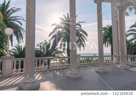 Sunlit terrace with columns and palm trees overlooking the ocean in a serene coastal setting 132185638
