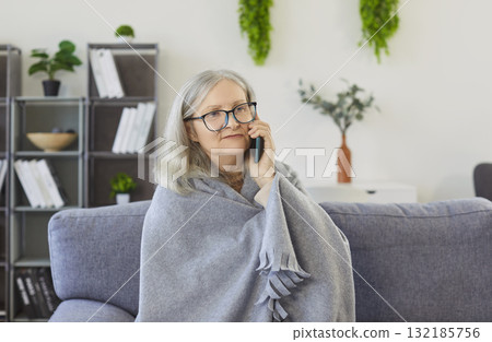 Serious senior woman wrapped in blanket sitting on sofa, talking on mobile phone and feeling unwell. Serious senior woman wrapped in blanket sitting on sofa, talking on mobile phone and feeling unwell. 132185756