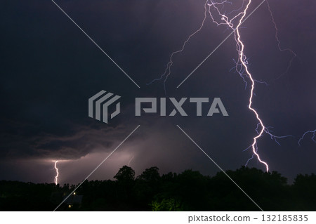 Night photo of two lightnings in the sky over a small village. One lightning close to the viewer, the other one smaller in the background 132185835
