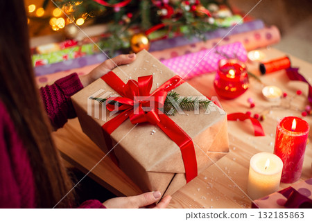 Girl checks present after wrapping it. Preparing gifts for friends and relatives in Christmas eve. Wrapping paper, candles, different decorations on the table Girl checks present after wrapping it. Preparing gifts for friends and relatives in Christmas eve. Wrapping paper, candles, different decorations on the table 132185863