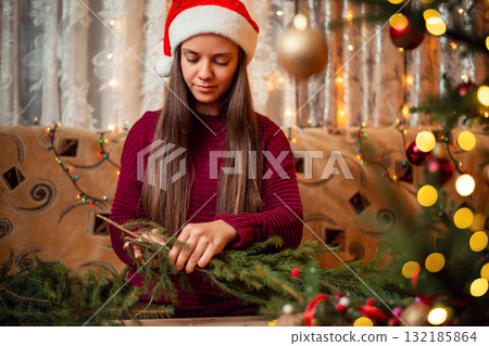 Portrait of a woman in red santa hat, sitting in sofa and making christmas wreath. Festive Christmas atmosphere, homemade decorations Portrait of a woman in red santa hat, sitting in sofa and making christmas wreath. Festive Christmas atmosphere, homemade decorations 132185864