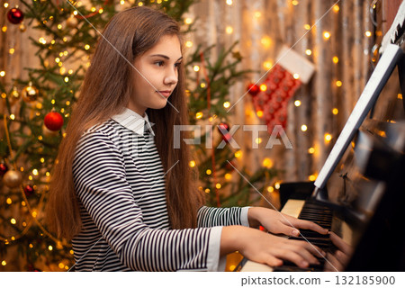 Young girl in a dress with long hair sitting near the piano and playing, Christmas tree on the background. Concept of the New Year, family holidays. Young girl in a dress with long hair sitting near the piano and playing, Christmas tree on the background. Concept of the New Year, family holidays. 132185900